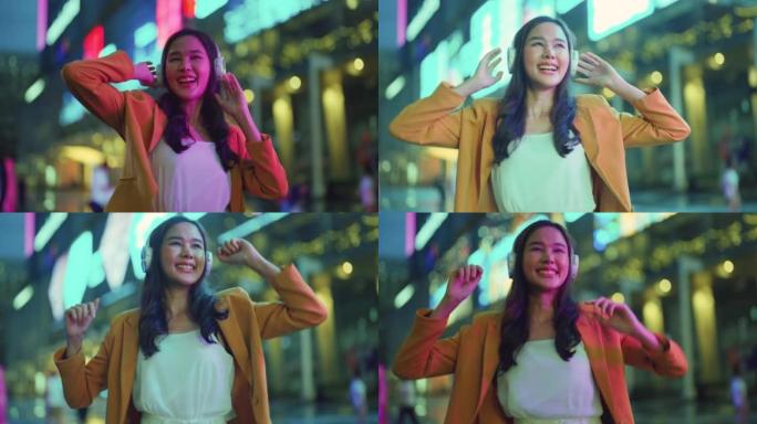 时尚着装的亚洲年轻女子感受快乐无忧无虑的耳机跳舞听音乐歌曲，快乐的女人跳舞自由移动在霓虹灯照明建筑的