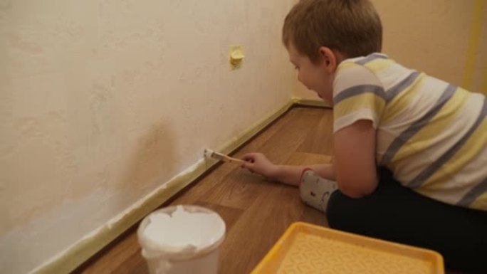 高加索男孩坐在地板上，在踢脚板附近用刷子粉刷墙壁