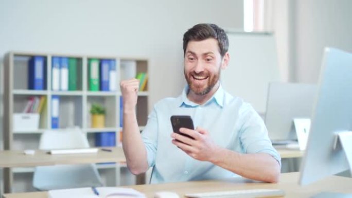 成功的帅哥员工通过在智能手机上查看消息来庆祝胜利，在移动应用程序中享受大奖坐在办公室