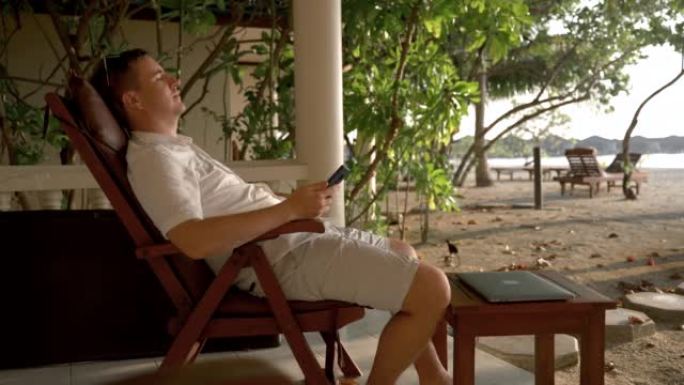 一名白人男子放下手机，在沙滩上的椅子上休息