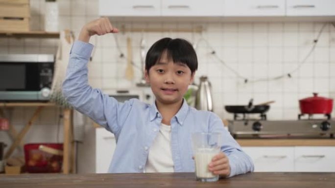 可爱的小微笑东亚顽皮的男孩拿着或喝着装满牛奶的杯子，在厨房背景下举起手臂