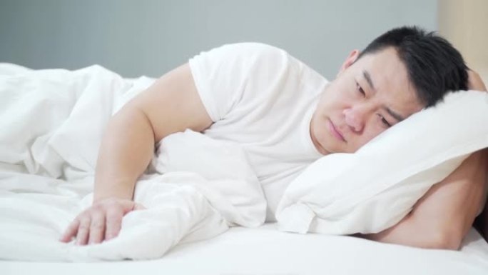 年轻的亚洲男子躺在床上悲伤与抑郁在家里的卧室。感觉不好。郁郁寡欢、精疲力竭、深思熟虑的男人