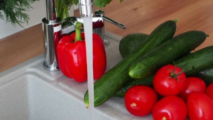 在水槽旁边的桌子上的自来水下洗的蔬菜。黄瓜，西红柿，甜椒，香草。相机移动变焦。健康饮食、素食主义的概