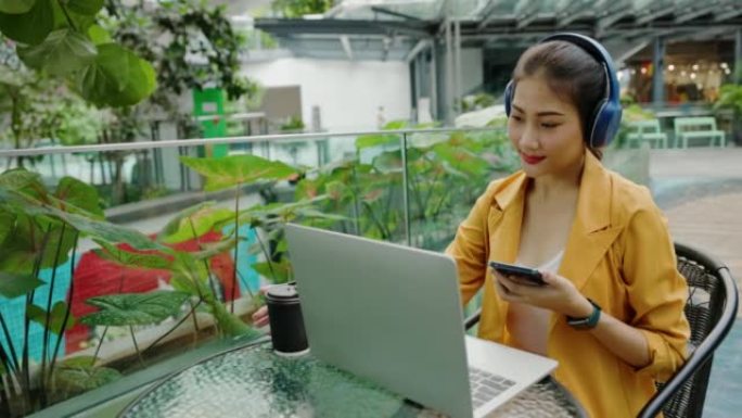 亚洲女商人坐在蓝牙耳机在户外笔记本电脑上工作快乐的微笑。女士快乐地使用平板电脑公园。利用现代互联网通