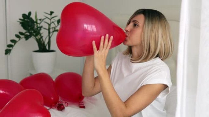 兴奋的女孩在床上给红心气球充气。女人在情人节欢喜礼物。随和的金发女孩在情人节表达积极的情绪。