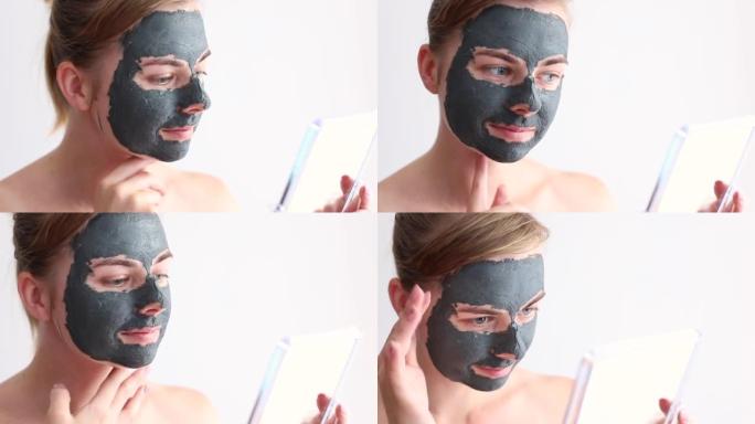 一个戴着黑色粘土化妆品面具的女人照镜子。在家中的美容护理