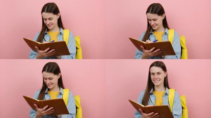 迷人的少女学生肖像在练习册笔记中写下，穿着衬衫和黄色背包，在粉红色背景工作室中孤立地摆姿势。大学学院