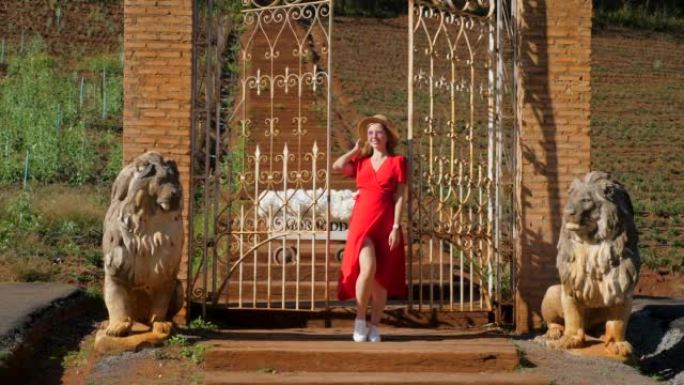 迷人，时尚的女人穿着红色连衣裙，在日落灯光下的殖民风格公园里，在锻铁大门旁摆姿势，狮子雕塑。时尚概念