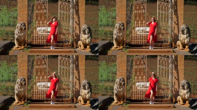 迷人，时尚的女人穿着红色连衣裙，在日落灯光下的殖民风格公园里，在锻铁大门旁摆姿势，狮子雕塑。时尚概念