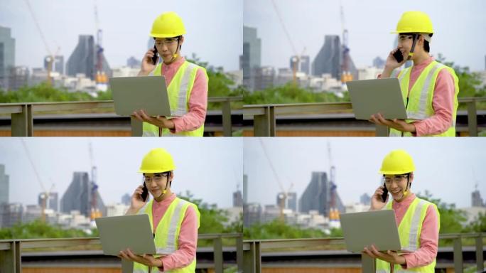 穿着安全帽和安全背心的快乐亚洲男性建筑工地经理在未完成的高层建筑内工作时在笔记本电脑上工作并在手机上