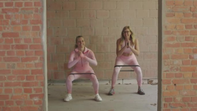 两名穿着粉红色运动服装的白人妇女在室内用橡皮筋下蹲