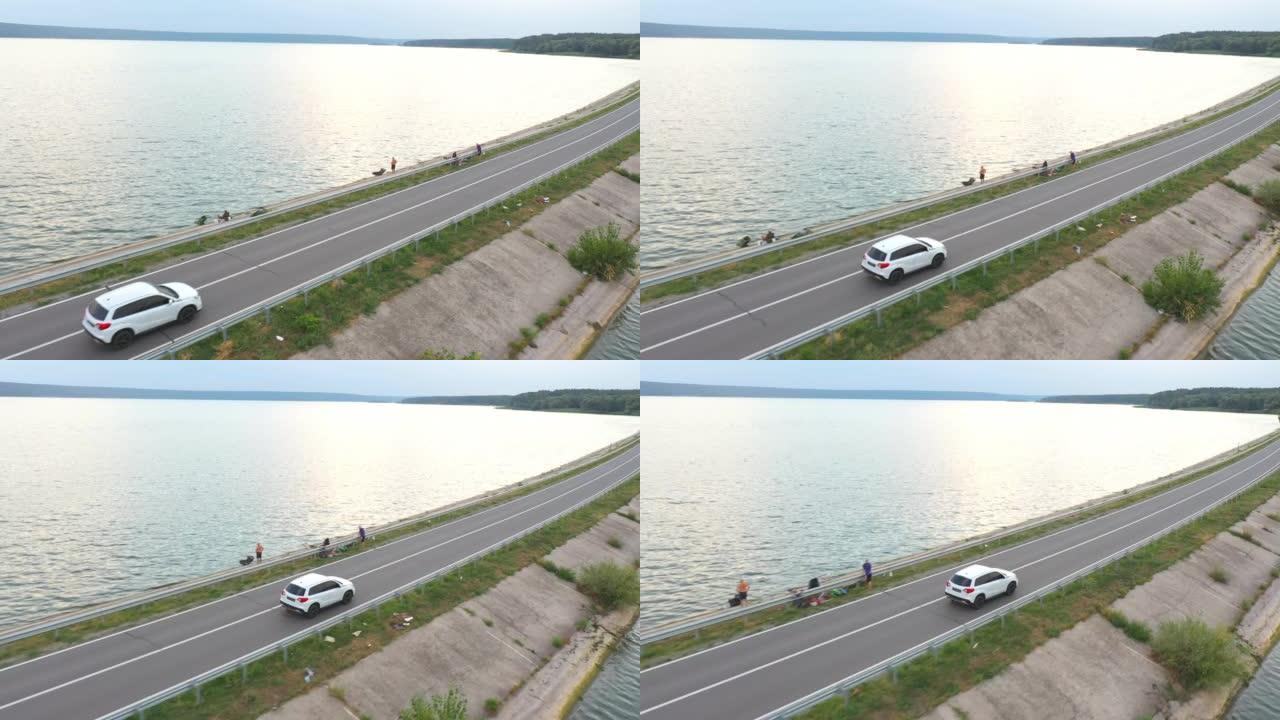 夏日白色汽车穿越大坝路线的空中拍摄。SUV在靠近湖泊或池塘的道路上行驶。飞越汽车穿过河桥。周末旅行或