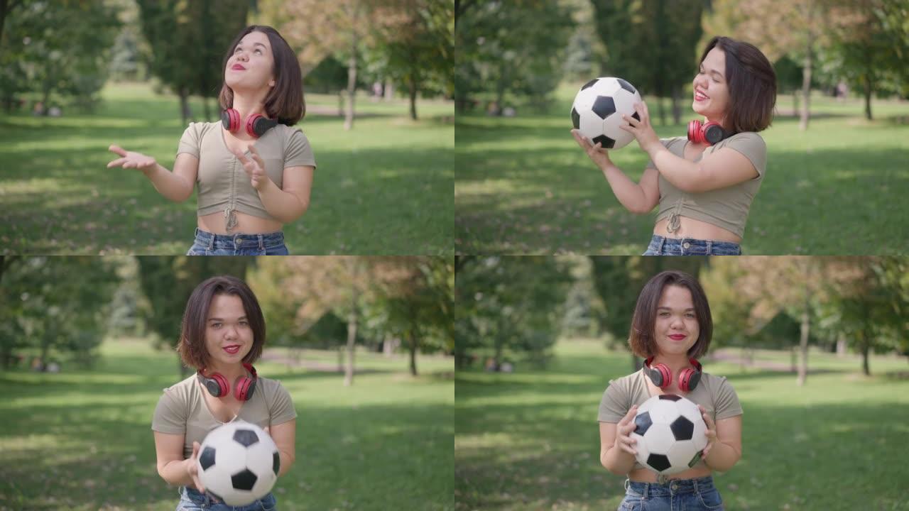 快乐健康的小女人在阳光明媚的公园里看着相机微笑着玩足球。自信的积极的白人侏儒症年轻人在户外慢动作摆姿