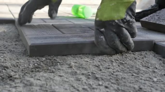 建筑商的双手戴着工作手套，将铺路石放在均匀的沙子上，并用沉重的塑料锤敲打瓷砖。慢动作，细节