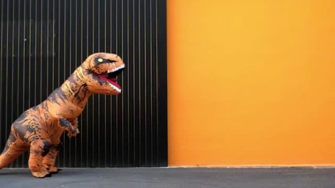 一件快乐而有趣的恐龙服装在街上跳舞，背景是橙色的彩色 -- 霸王龙在恐龙服装里玩得很开心 -- 有趣