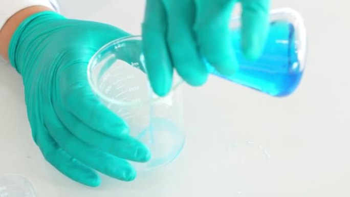 专业化学家的特写手穿着防护制服，戴着手套，将移液器中的溶液加入到烧瓶中的蓝色混合物中。在现代实验室工