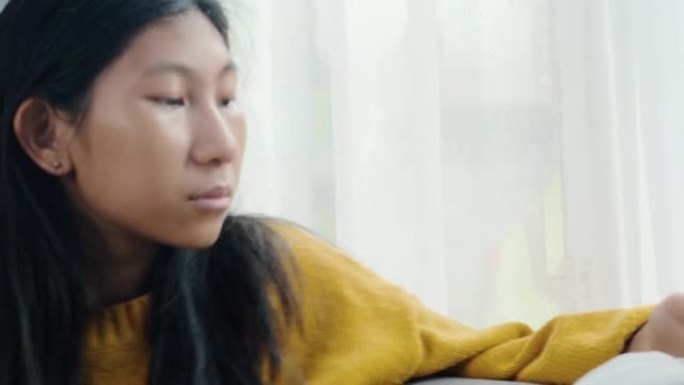 在雨天，穿着黄色毛衣的亚洲女孩在窗户附近的沙发上使用智能手机，生活方式概念。