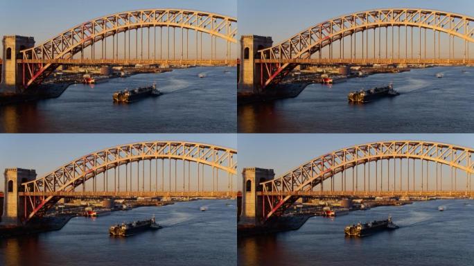 一艘驳船在纽约，长岛市，皇后区的地狱门桥下的东河上行驶，在停泊船只的商业文件前。航空无人机制作的带静