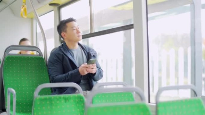 亚洲男子乘坐公共交通工具，坐在座位上使用手机，智能手机在公交车窗前。城市背景上的男性休闲肖像