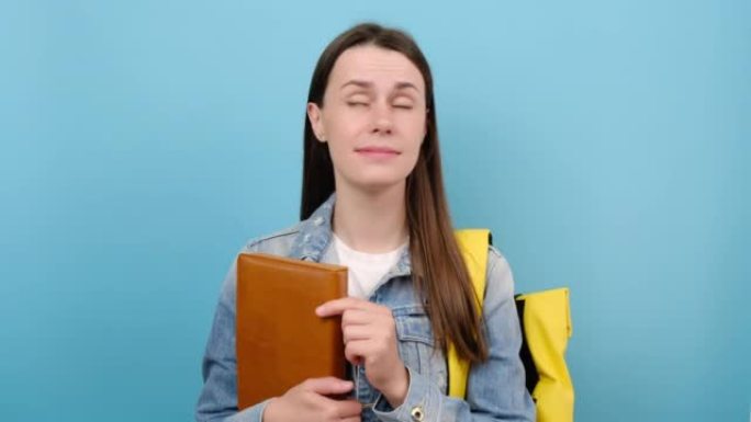 少女学生的肖像穿着牛仔夹克和黄色背包拿着书说哎呀哎呀天哪，在蓝色背景上孤立地摆姿势。高中教育大学学院