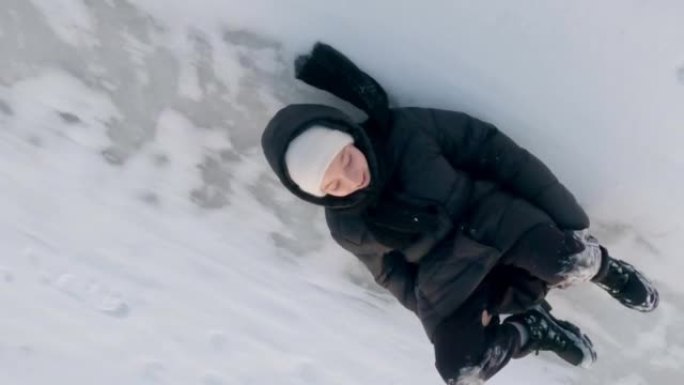 穿着黑色衣服的快乐少女在冬天霜冻的日子里，没有雪橇的雪冰滑梯在后面滑下来，躺在雪地上。冬季运动会。