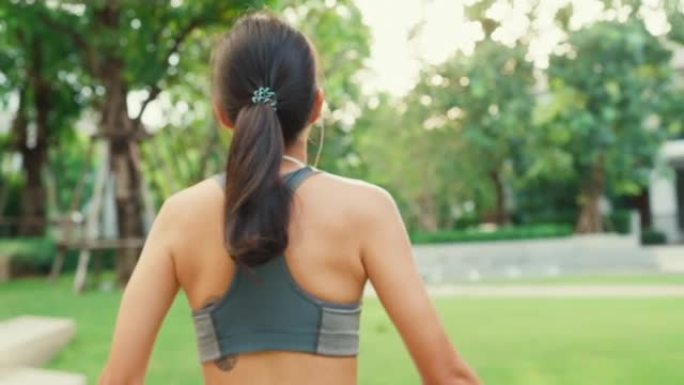 特写快乐的年轻运动型亚洲女性穿着带耳机的运动服，从后角度听音乐，从后角度散步和伸展放松，在花园公园锻