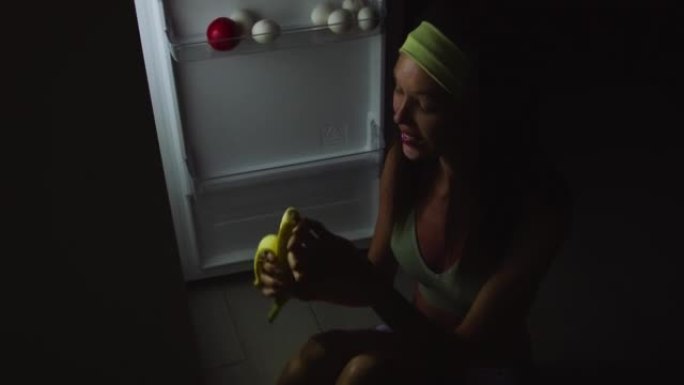 一个年轻饥饿的女人的特写镜头在晚上打开冰箱，坐在地板上吃东西。