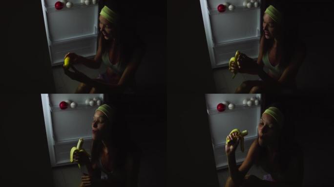 一个年轻饥饿的女人的特写镜头在晚上打开冰箱，坐在地板上吃东西。
