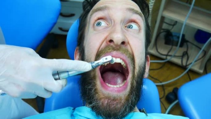 一个年轻的大胡子男人害怕专业人士刷牙，坐在牙医的椅子上惊恐地尖叫
