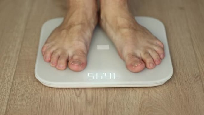 步行男性检查BMI体重减轻。人体赤脚测量身体脂肪超重。男人的腿踩在浴室秤上。减肥人的脚站在房间里称重