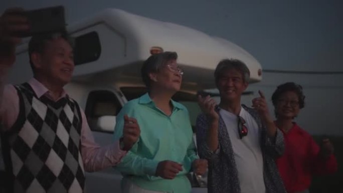 日落时，亚洲华裔高级男子在稻田露营车旁使用智能手机与他的高级朋友唱歌和自拍肖像