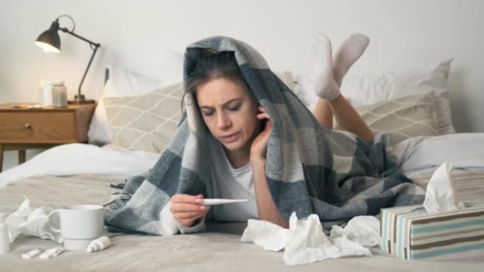 患有流感的不健康妇女躺在床上看温度计上的温度。发热高的卧室里生病的女人。感冒和偏头痛