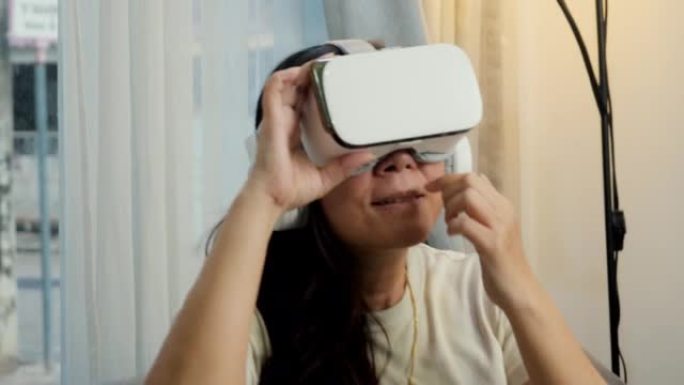 亚洲妈妈在家靠窗的沙发上通过虚拟现实玩游戏，科技生活理念。