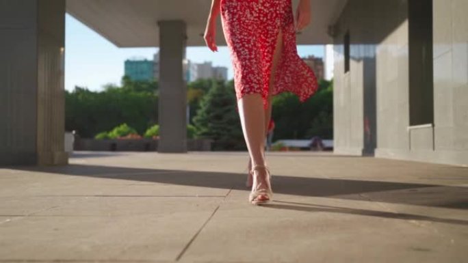 穿着高跟凉鞋的苗条女性腿和红色连衣裙沿着城市阳光明媚的街道漫步。美丽的女孩在市中心的夏天走来走去。面