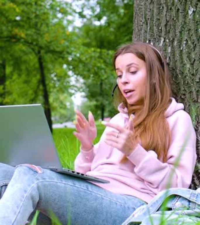 垂直视频-坐在城市公园时，戴着笔记本电脑和耳机的年轻女子进行在线对话。