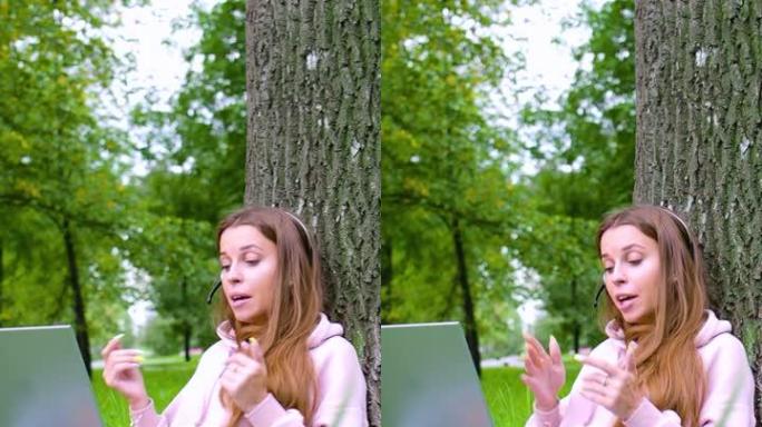 垂直视频-坐在城市公园时，戴着笔记本电脑和耳机的年轻女子进行在线对话。