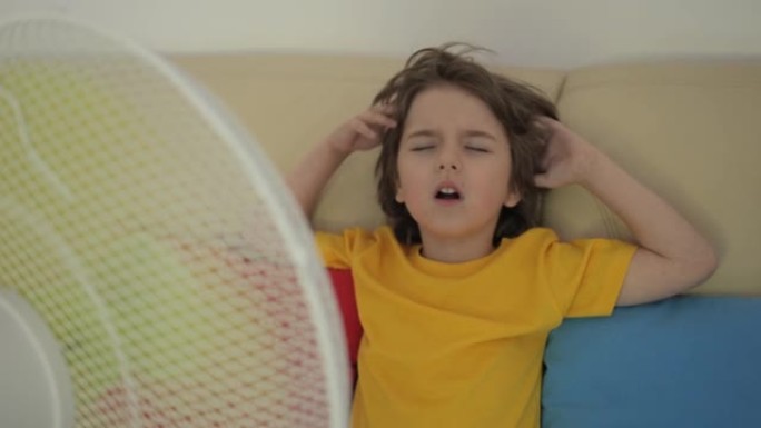 男孩坐在风扇热天的沙发上。夏天炎热时，孩子在家享受电风扇带来的凉风。使用电风扇冷却器冷却自己的通风机