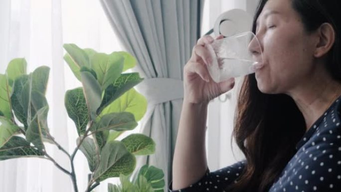 亚洲成年妇女在家中窗户附近喝一杯凉水，简单的生活方式概念。