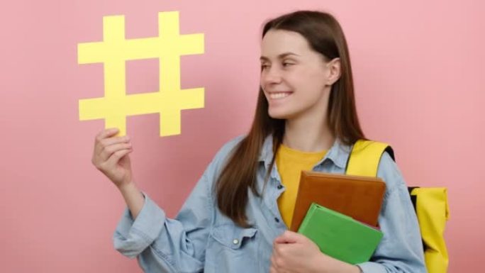 快乐美丽女孩的肖像青少年学生穿着背包拿着书，显示黄色标签，孤立在工作室的粉色背景墙上，有复制空间。概