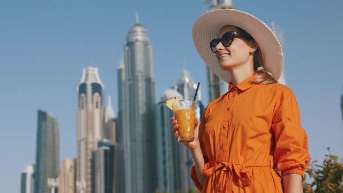 迪拜旅游。城市天际线背景上穿着橙色连衣裙和太阳帽搭配冰沙鸡尾酒的旅游女性。阿联酋的假期