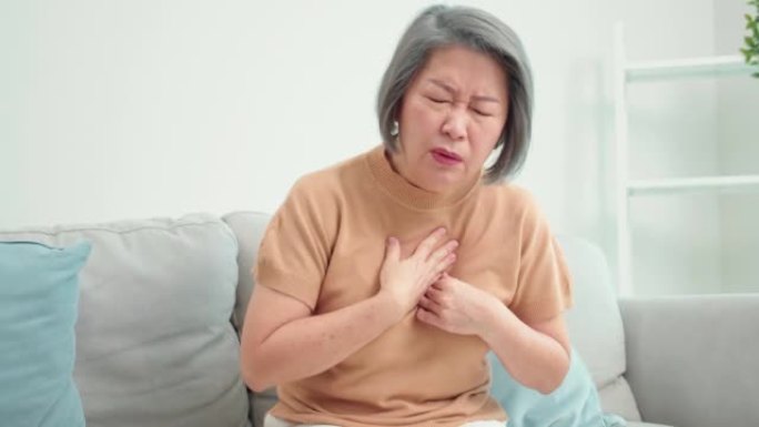 亚洲老年老年亚洲妇女心脏病发作，坐在家里客厅的沙发上。患有痛苦和胸痛的老年女性因急性疼痛而难以呼吸。