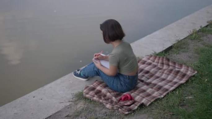坐在夏日公园湖岸的体贴高加索小女人写日记的高角度视图。宽镜头快乐放松的侏儒症年轻人梦想在户外休息。