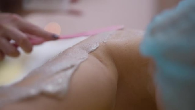 用刮刀覆盖客户皮肤的特写手。无法辨认的高加索美容师在美容水疗中心的裸体女人的肩膀上涂抹嫩肤化妆品。慢