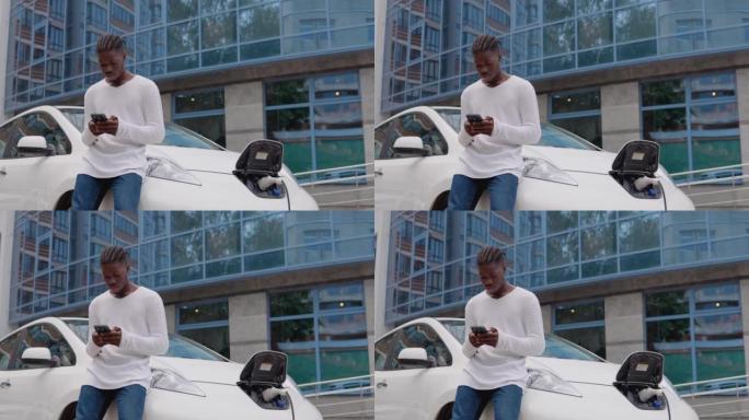时尚的年轻非洲裔美国男子站在电动汽车附近，用智能手机充电和刷卡