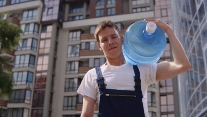 Courier在现代高层建筑的背景下，肩上扛着一大罐饮用水，看着镜头。送水回家