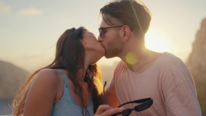 浪漫的夏天。夫妇在度假时戴着墨镜坐在长凳上，背景是金色的日落。热情温柔的吻