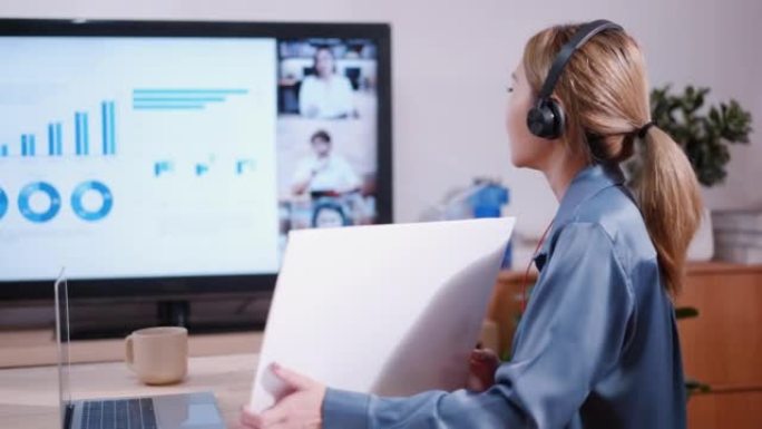 年轻的亚洲女商人戴着耳机坐在她的桌子上，用笔记本电脑和电脑屏幕在网上会议上向她的同事展示商业计划表
