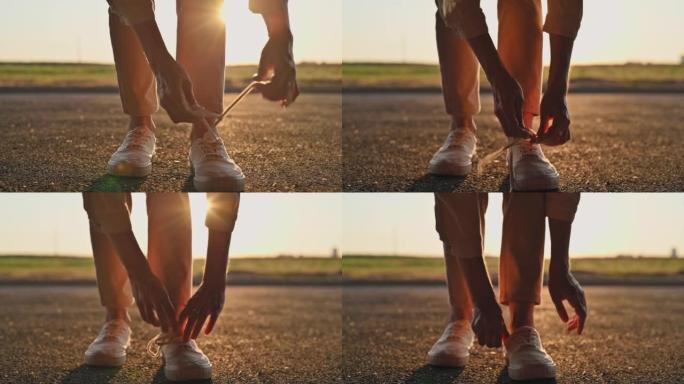 年轻迷人的女孩穿着米色牛仔裤，在日落时分的城市道路上系上白色运动鞋的蕾丝特写镜头。带有夕阳光线的电影