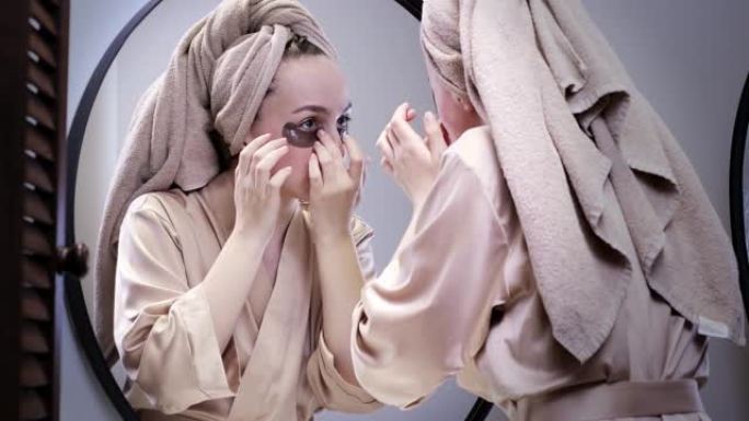一位穿着浴袍和毛巾的穿着整齐的女人在家中因肿胀和瘀伤而在眼睛下方涂抹补丁。制作美容程序-在眼睛下涂抹
