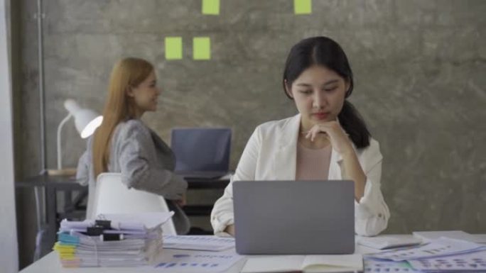 两名年轻的亚洲女商人负责财务文件，分析投资项目和规划策略的信息。商人与笔记本电脑交谈，在办公室在线工